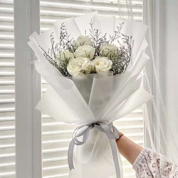 Elegant bouquet of white roses with delicate greenery, wrapped in white paper and tied with a gray ribbon.