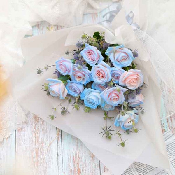 Bouquet of light blue and pink roses with greenery, wrapped in white paper.