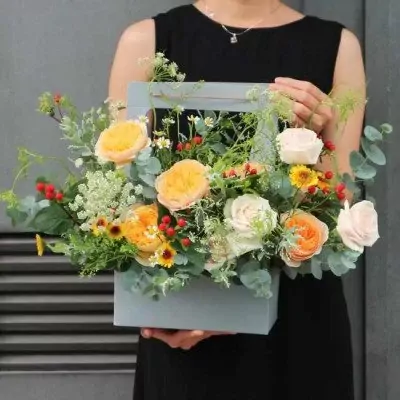 A woman in a black dress holds a pale blue floral arrangement box with various flowers including yellow and pink roses, small yellow sunflowers, and red berries.