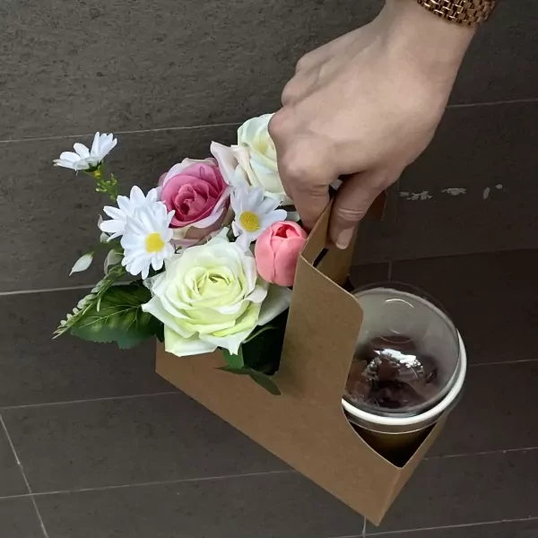 Hand holding a brown cardboard carrier with a bouquet of artificial roses, daisies, and tulips, and a cup of chocolate drink.