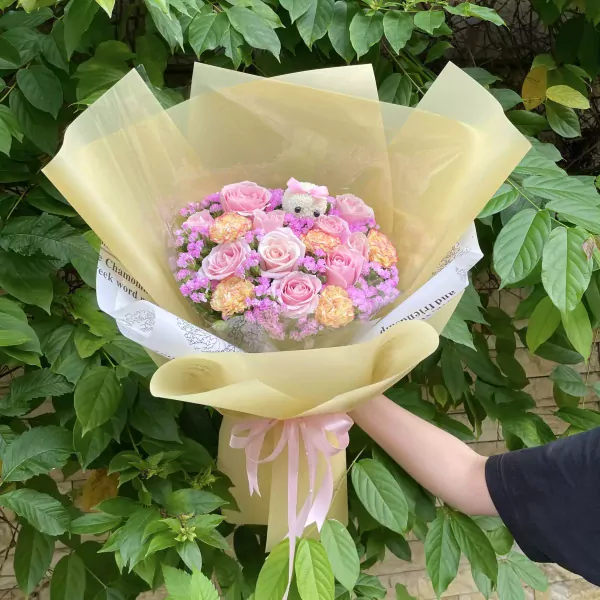 Pink and cream roses, carnations, and baby's breath bouquet with a teddy bear, wrapped in yellow paper.