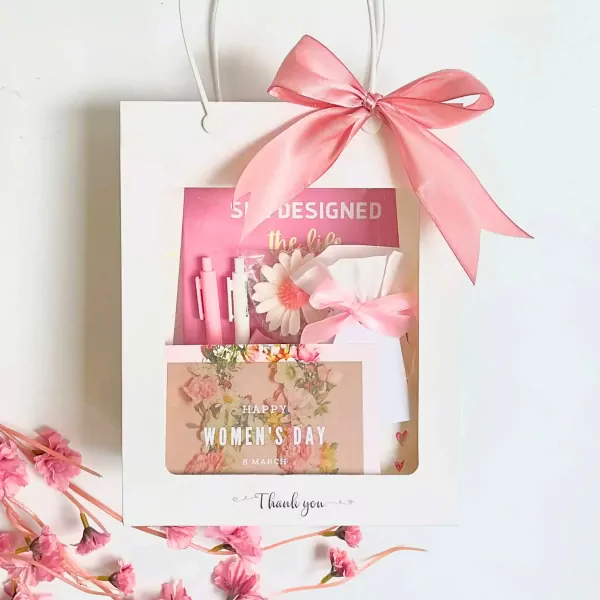 Pink gift bag with a ribbon bow, featuring pens, a notebook, a flower, and a Women's Day card.