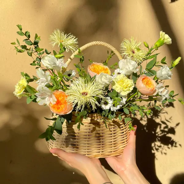 Woven basket overflowing with vibrant orange, yellow, and white flowers, held by two hands.