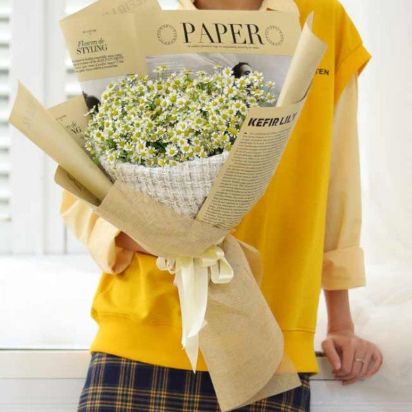 A hand holding a bouquet of white daisies wrapped in newspaper-style paper and a beige tweed fabric.