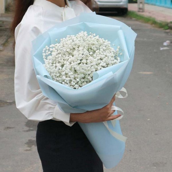 A woman in a white blouse holds a bouquet of white baby's breath flowers wrapped in light blue paper.