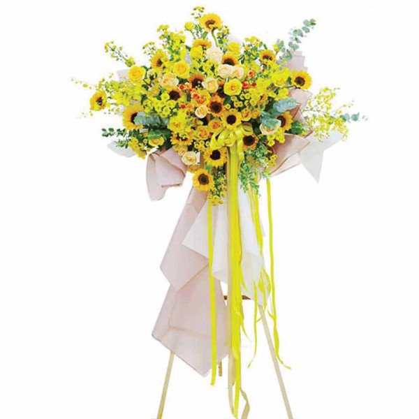 Yellow flower arrangement with sunflowers, roses, and orchids on a stand with pink and yellow ribbons.