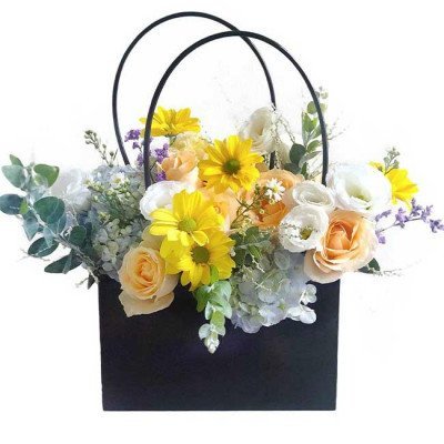 A black gift bag filled with a mixed flower arrangement featuring yellow daisies, peach roses, white roses, and blue hydrangea.