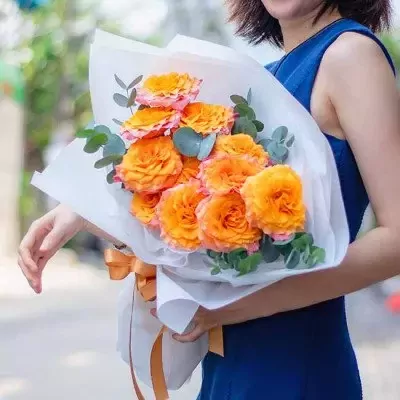 Woman holding a bouquet of vibrant orange and pink roses wrapped in white paper and tied with an orange ribbon.