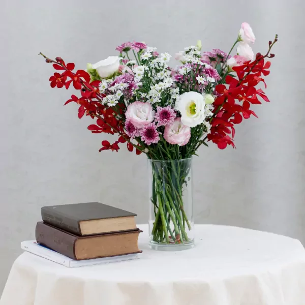 A bouquet of red orchids, purple mums, and pink lisianthus in a glass vase on a table with books.