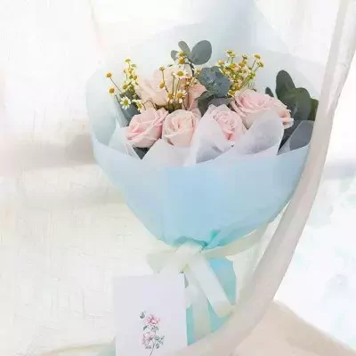 Bouquet of pink roses, chamomile, and eucalyptus leaves wrapped in pale blue paper with a white ribbon and card.