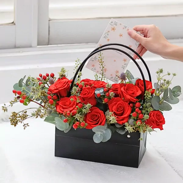 A hand places a floral card into a black box filled with red roses, hypericum berries, and eucalyptus.