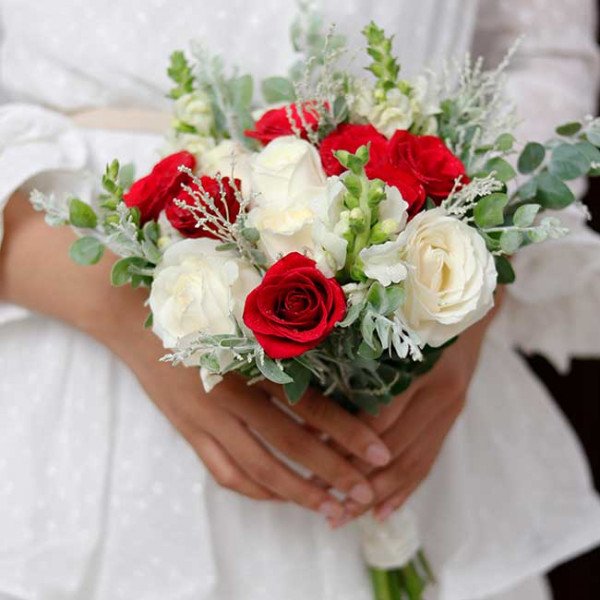 Bridal bouquet of red and white roses with greenery held by a person in a white dress.