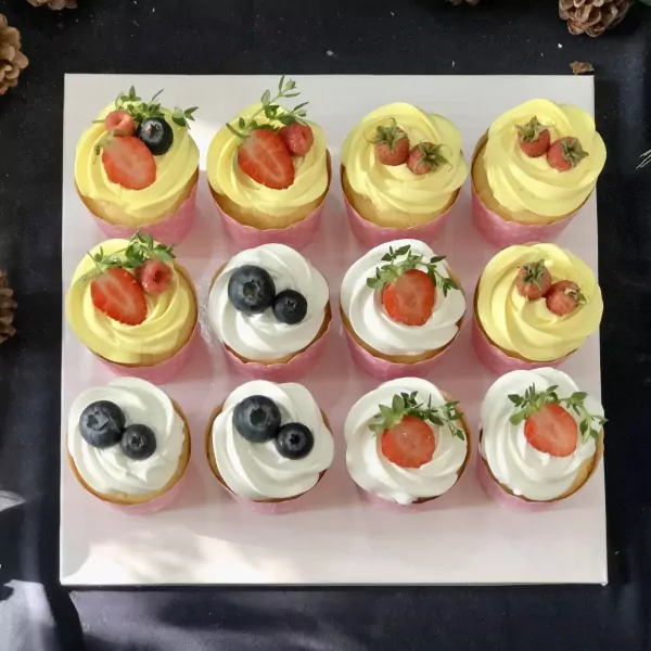 Assortment of 12 cupcakes with yellow and white frosting, topped with strawberries and blueberries.