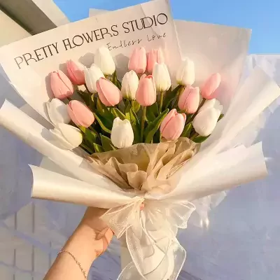 Hand holding a bouquet of pink and white tulips wrapped in white and brown paper with a sheer ribbon.