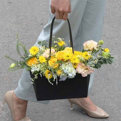Woman in pale blue pants and nude heels carries a black basket of yellow roses, hydrangeas, and carnations.