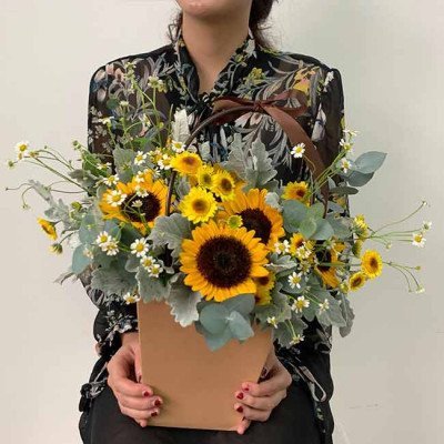 Woman holding a basket of sunflowers, daisies, and greenery with a brown ribbon.