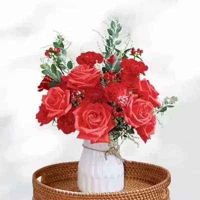 Red roses and carnations in a white vase on a wicker tray.