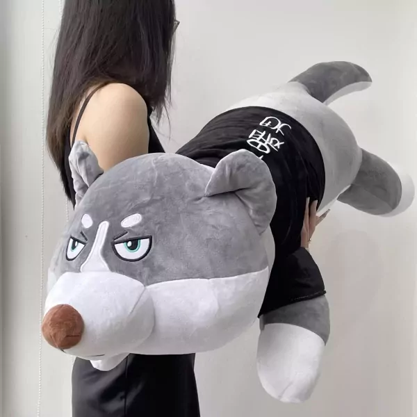 A person holds a large gray husky plush toy wearing a black shirt with white lettering.