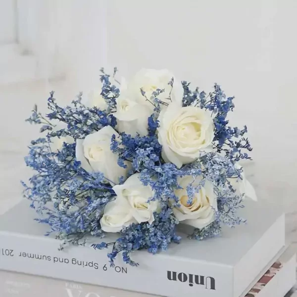 White roses and blue filler flowers arranged in a bridal bouquet on top of books.