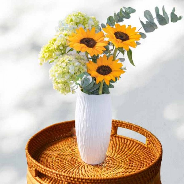 Bouquet of sunflowers and hydrangeas in a white vase on a rattan tray.