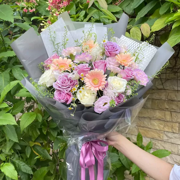 Violet and pink roses, white roses, gerbera daisies, and carnations in a bouquet with grey and white wrapping.