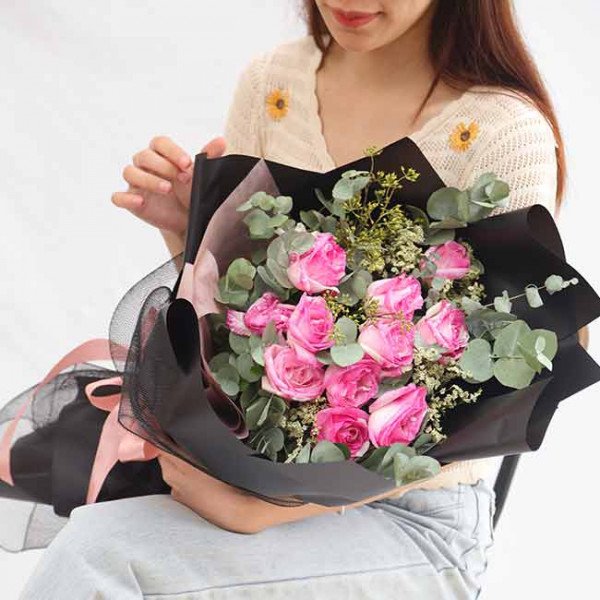 Woman holding a bouquet of pink roses and eucalyptus leaves wrapped in black paper.