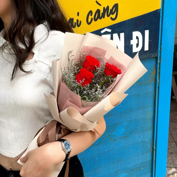 Three red roses in a bouquet wrapped in paper, held by a person wearing a white top and watch.