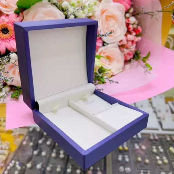A purple velvet jewelry box with a pearl necklace nestled inside, with a bouquet of pink and white flowers in the background.