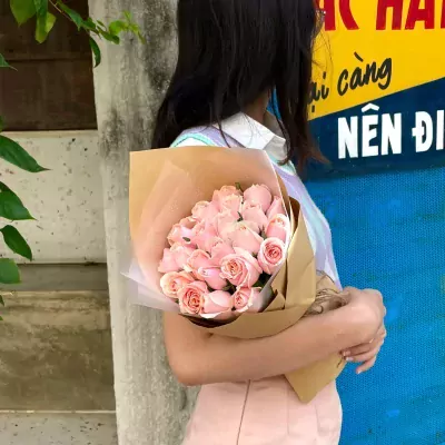A bouquet of pink roses wrapped in brown paper, held by a person in a pastel sweater and pink skirt.