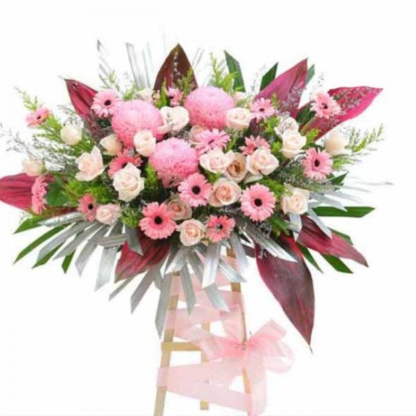 Pink floral arrangement with roses, carnations, and gerbera daisies on a wooden stand.