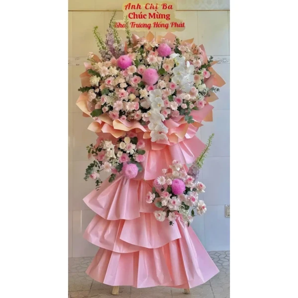 A pink tiered floral arrangement with roses, orchids, and other flowers on a stand with a congratulatory banner.