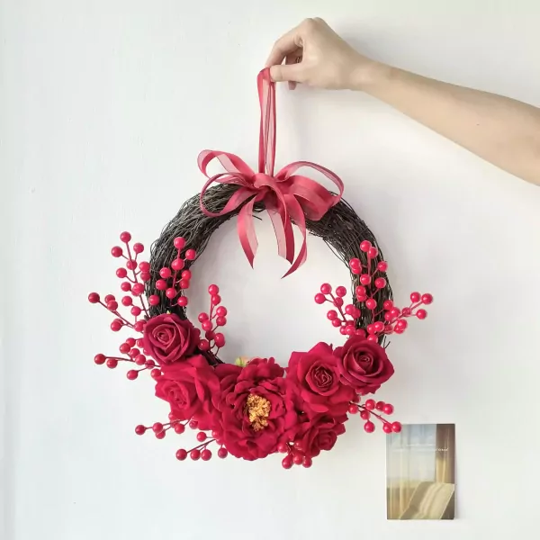 Red floral wreath with berries and a bow hanging from a ribbon on a white wall.