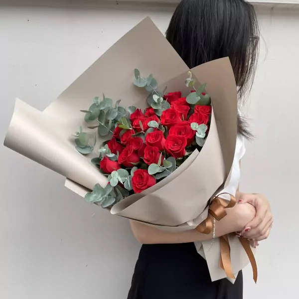 A bouquet of 24 red roses with eucalyptus, wrapped in beige paper and tied with a brown ribbon.