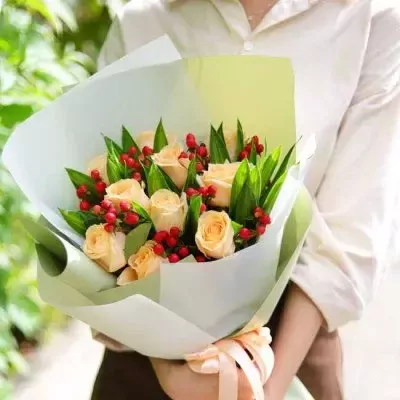 Bouquet of peach roses with red berries and green leaves, wrapped in white and green paper, tied with a peach ribbon.