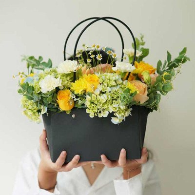 A black flower basket filled with yellow, white, and peach flowers, held by a person.
