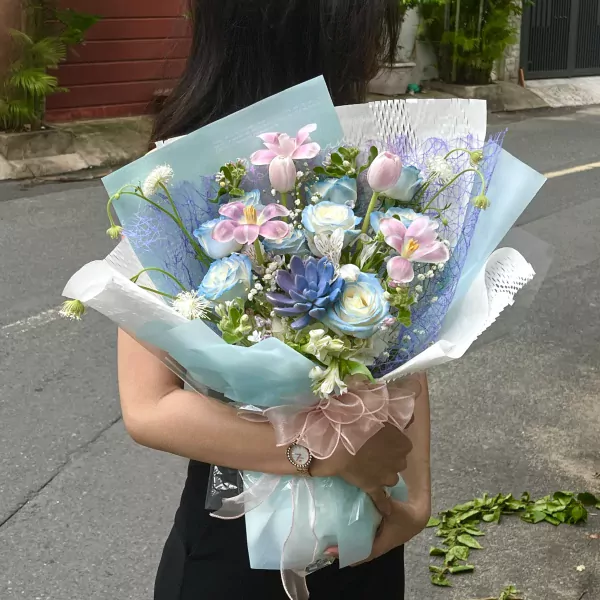 A woman holds a pastel pink and blue flower bouquet with roses, tulips, and succulents.