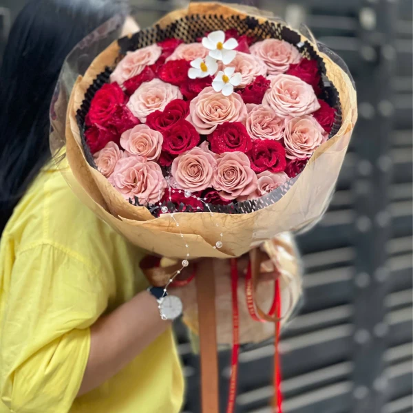 A woman in a yellow shirt holds a large bouquet of pink and red roses with small white flowers at the top.