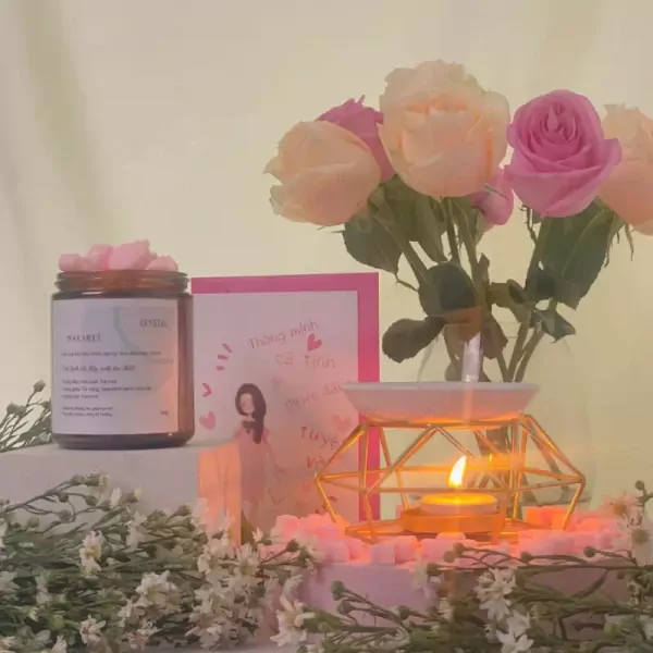 A jar of pink wax melts, a vase of pink and cream roses, and a lit oil diffuser on a pink display.