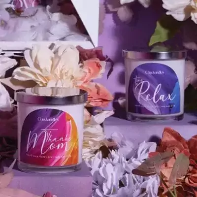 Two scented candles, one labeled "Thanks Mom" and the other "To Relax," surrounded by flowers.