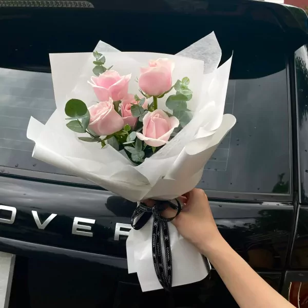 Bouquet of pink roses and eucalyptus, wrapped in white paper with a Chanel ribbon, held by a hand in front of a black car.