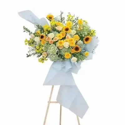 A large, bright floral arrangement with yellow roses, sunflowers, and hydrangeas on a wooden stand.