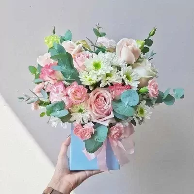 A hand holds a light blue box filled with pink and white roses, chrysanthemums, and eucalyptus leaves.