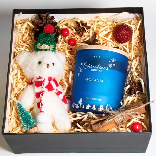 Christmas gift box with white teddy bear, blue scented candle, pine cone, and cinnamon sticks.