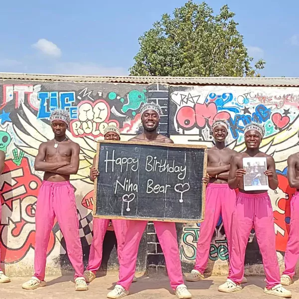 African men in pink pants and crowns hold a birthday sign and a photo.