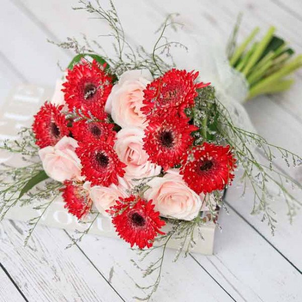 Bouquet of red gerbera daisies and pink roses, accented with delicate greenery, on a white wooden surface.
