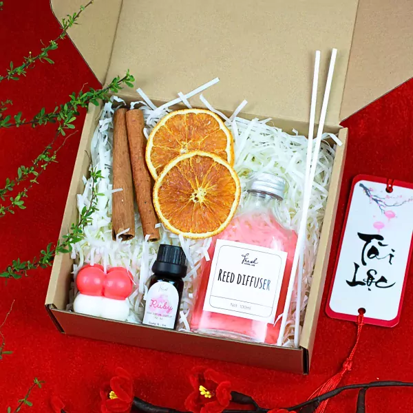 Gift box with red candle, reed diffuser, dried oranges, cinnamon sticks, and a tag.