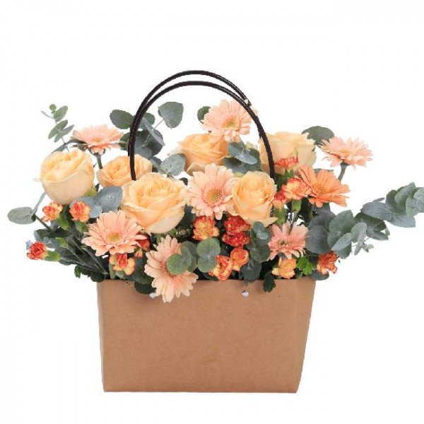 A flower basket with peach roses, gerbera daisies, and carnations in a brown paper bag with handles.