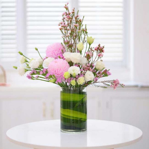 A floral arrangement in a clear glass vase with green accents. Features pink peonies, white roses, and delicate pink wildflowers.