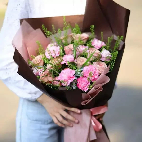 A bouquet of pink roses and carnations wrapped in brown and pink paper, held by a person in a white top and blue jeans.