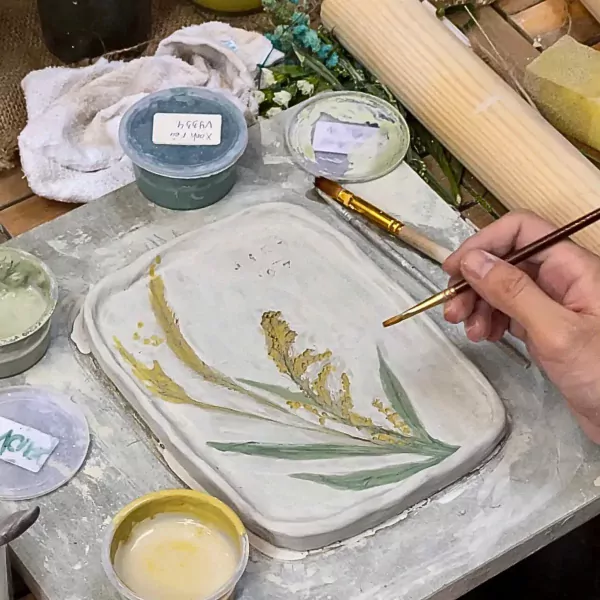 Hand painting a ceramic tray with wheat stalks and leaves at a pottery workshop.